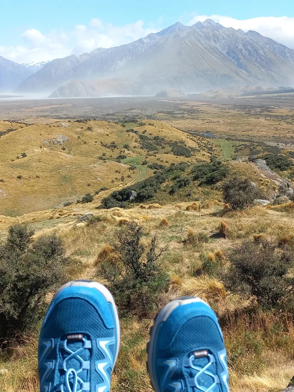 Judith's bergschoenen bungelend over een rots met uitzicht over het uitgestrekte landschap van Hakatere Conservation Park, Nieuw-Zeeland