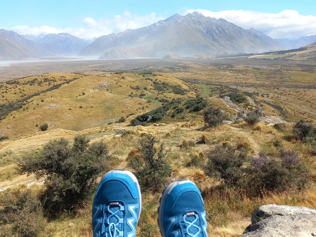 Judith's bergschoenen bungelend over een rots met uitzicht over het uitgestrekte landschap van Hakatere Conservation Park, Nieuw-Zeeland